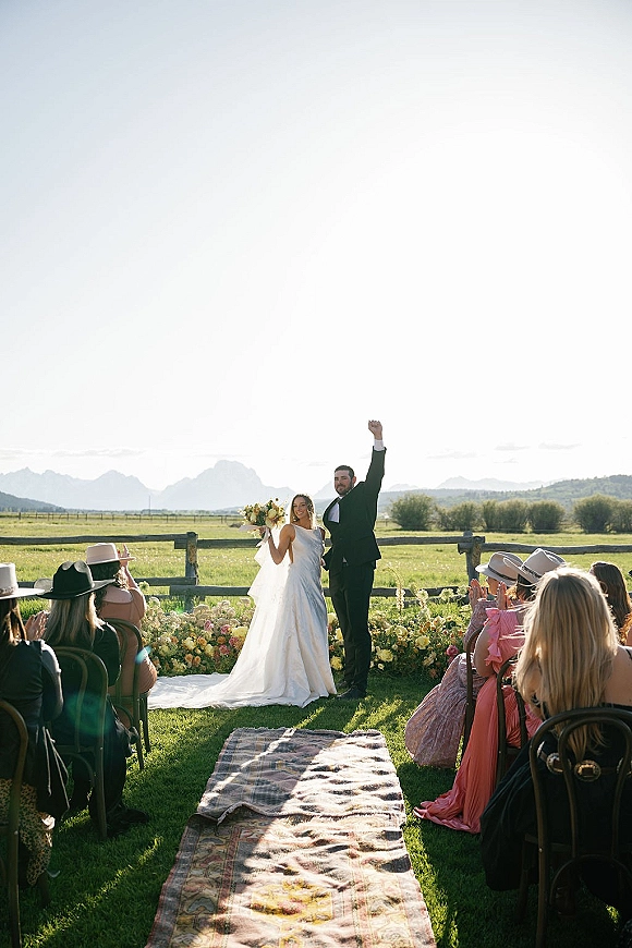 Recessional moment as bride and groom cheer walking down a vintage rug aisle runner, bride holding bouquet, mountains behind them