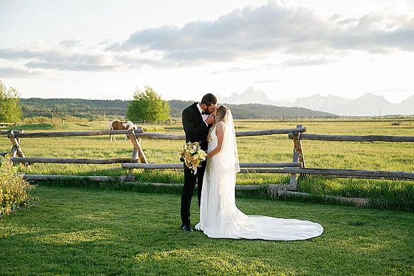 Wedding kiss portrait of bride and groom kissing, her veil and bouquet in a grassy field by a rustic fence with a horse and mountains under clouds