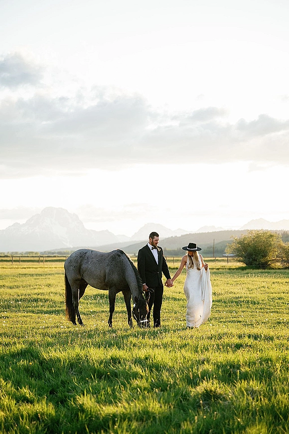 Couple portrait of bride in a wedding dress and hat holding hands with groom in black suit beside a horse in a mountain field under clouds