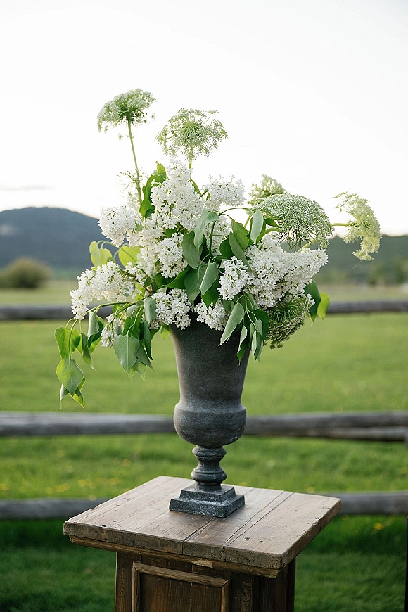 Wedding floral arrangement of white flowers and greenery in a black urn on a rustic wooden table, set in a mountain field backdrop