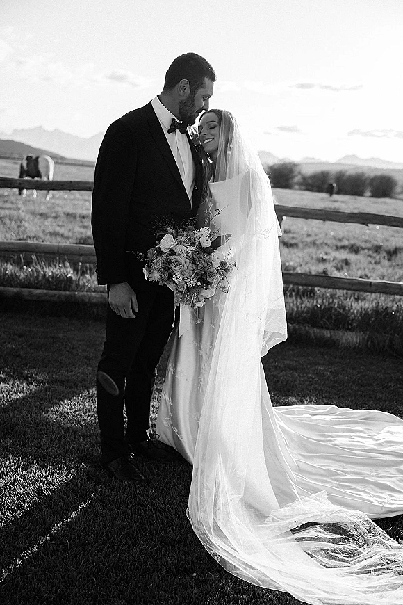 Couple portrait in a black and white wedding portrait, bride in long veil train holding bouquet as groom touches foreheads, mountains behind