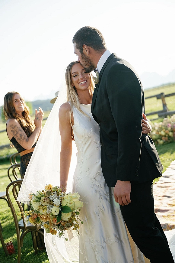 Wedding couple portrait of groom kissing bride’s forehead as she holds a bouquet, veil flowing on a mountain ceremony aisle with chairs