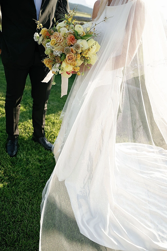 Wedding couple portrait of bride and groom standing on a grassy hill, bride holding a yellow bouquet with veil and dress train flowing in sky light