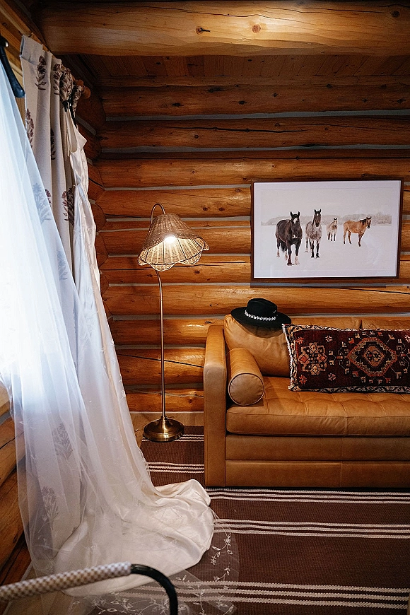 Wedding dress hanging by window, bridal dress by window with long train on woven rug in a log cabin room with sheer curtains