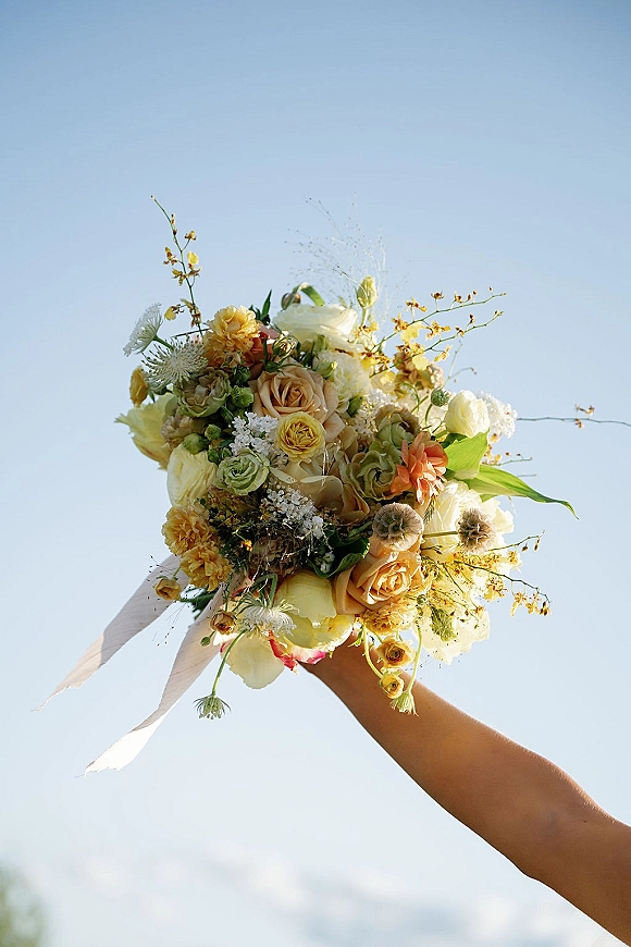 Bridal bouquet with yellow wedding bouquet tones, garden roses and ranunculus, tied with flowing ribbon against a blue sky backdrop