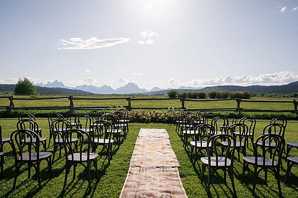 Outdoor ceremony setup with a wedding aisle runner on grass, wood chairs in rows, and floral ground arrangement facing mountains under clouds