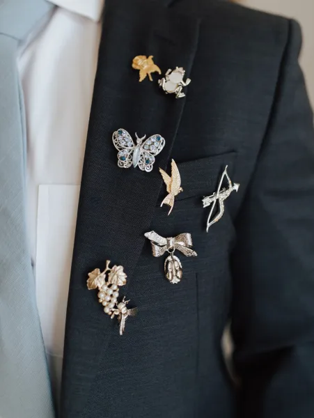 Groom lapel pins displayed on a suit jacket, featuring crystal butterfly and bird brooches with gold and silver details indoors