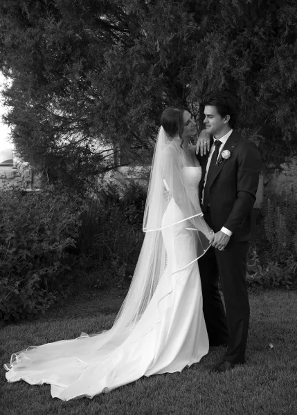 Couple portrait of a bride and groom holding hands on a grass lawn framed by trees and shrubs, her long veil trailing softly behind.