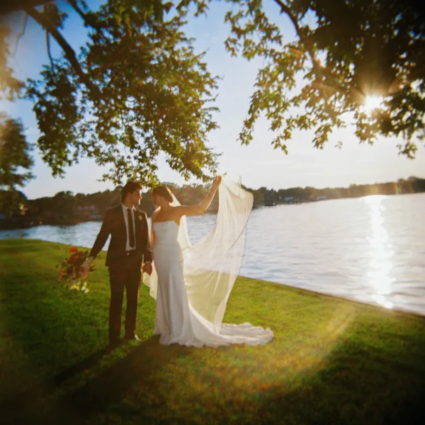 Couple portrait of a bride and groom holding hands by a sunlit lake, her bridal veil catching light beneath leafy tree branches.
