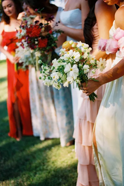 Bridesmaids photo with mismatched bridesmaid dresses as they stand on a sunlit lawn, holding hand-tied bouquets amid outdoor greenery.
