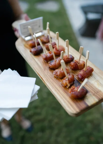 Wedding appetizers arranged as passed hors d'oeuvres on a wooden serving board, ready for guests on a grass lawn.