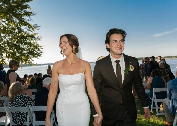 Wedding recessional with a couple walking down the aisle hand in hand beside a shimmering lake, the bride’s veil flowing under a clear blue sky.