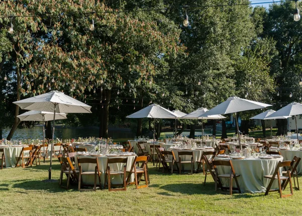 Outdoor reception setup with garden reception decor, round tables under patio umbrellas arranged on a grassy lawn beside a tranquil lake.