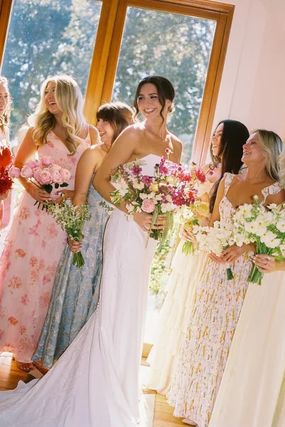 Bride and bridesmaids in mismatched bridesmaid dresses pose with garden-style bouquets by a large window framing leafy trees.