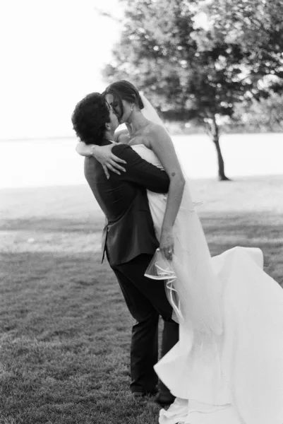 Wedding kiss portrait with the groom lifting the bride, her veil drifting, on a grassy lawn by the water framed with trees under open sky.