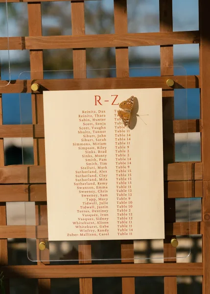 Wedding seating chart and table assignments sign mounted on a wood trellis, with a serene backdrop of water, trees, and open sky.