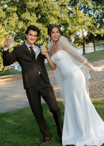 Couple portrait of a bride and groom celebrating with cocktails on a lakeside lawn, framed by garden trees and a winding gravel path.