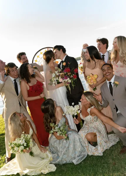 Wedding kiss portrait of the couple amid a cheering wedding party group photo on a lakeside lawn, the bride’s veil catching the light.