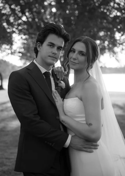 Couple portrait as the bride and groom embrace beside a lake, her veil flowing as he holds her waist amid trees on the lawn.