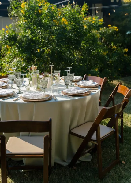 Reception tablescape for an outdoor wedding table set with taper candles, surrounded by garden trees and a lawn under string lights.