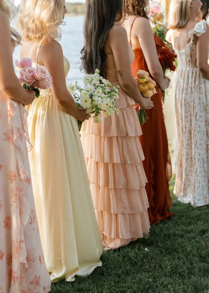 Bridesmaid dresses, including a yellow bridesmaid dress, in peach, rust, and floral prints with ruffled tiers on a grassy lakeside lawn.