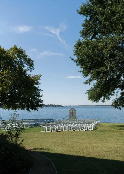 Outdoor ceremony setup for a lakeside wedding ceremony with a stained glass arch, framed by a calm lake, shoreline trees, and open blue sky.