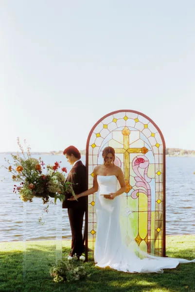 Private vow exchange during a first touch moment as the couple stands back to back, holding hands by a lakeside lawn with stained glass panel.