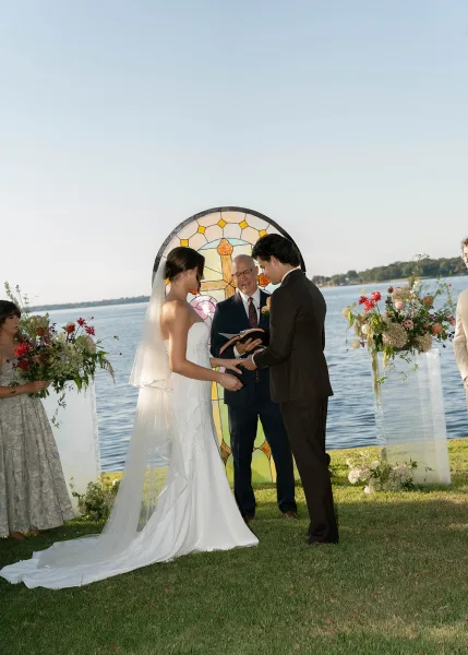 Ceremony moment during an outdoor wedding ceremony as the couple holds hands beneath a stained glass backdrop by the lake.