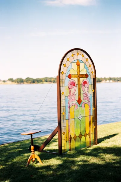 Ceremony backdrop with a stained glass altar set on a lakeside lawn, blue sky and distant trees reflecting off the calm water.