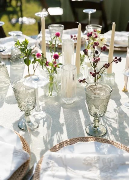 Reception tablescape for an outdoor reception table with taper candles on a grass lawn, flanked by wooden chairs.