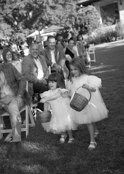 Flower girls in flower girl dresses hold flower baskets on a garden lawn near ceremony chairs and shady trees.