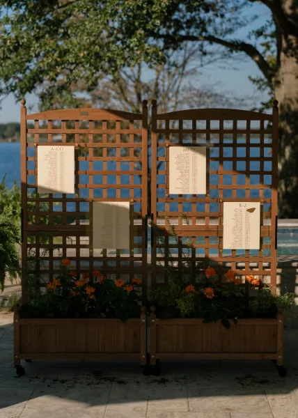 Seating chart display styled as a trellis seating chart with orange flowers, set by a lake and trees on a stone patio.