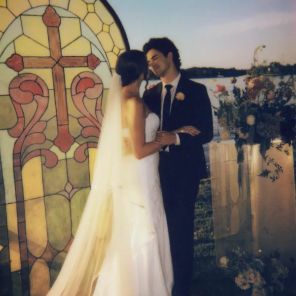 Couple portrait of a bride and groom holding hands on a lakeside lawn, her long veil trailing as they face each other under open sky.