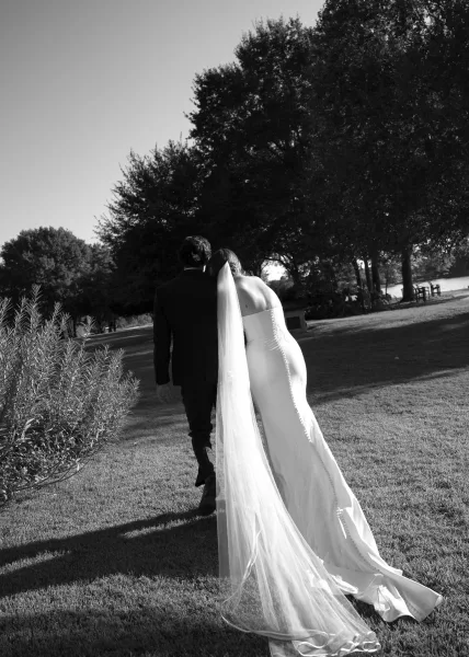 Bride and groom walking in a black and white wedding photo across a lakeside lawn, her long veil trailing as trees frame the sky.