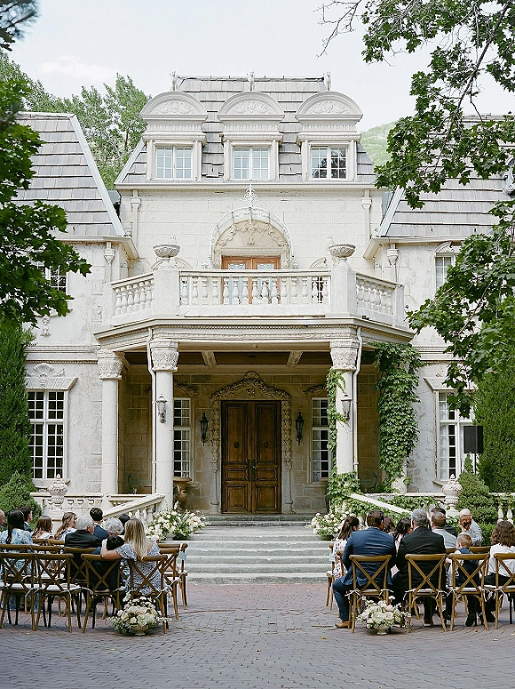 Ceremony setup with outdoor ceremony seating of cross back chairs lining an aisle with white florals on stone steps before ivy-covered manor doors