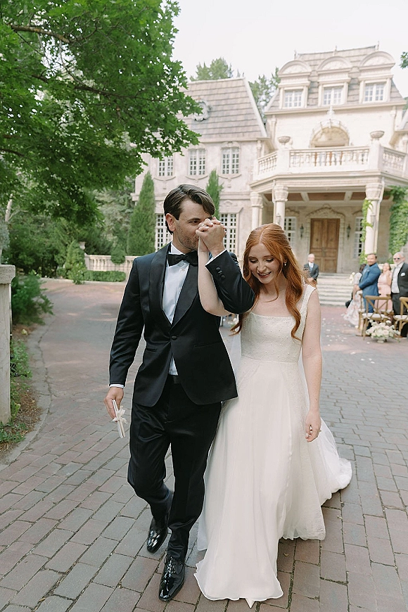 Wedding couple portrait of bride and groom walking hand in hand on a stone driveway by an estate house, bride in veil and satin gown