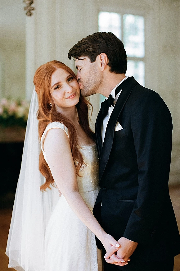 Wedding couple portrait of groom kissing bride’s forehead as they hold hands, her long veil and his black tuxedo in soft window light indoors