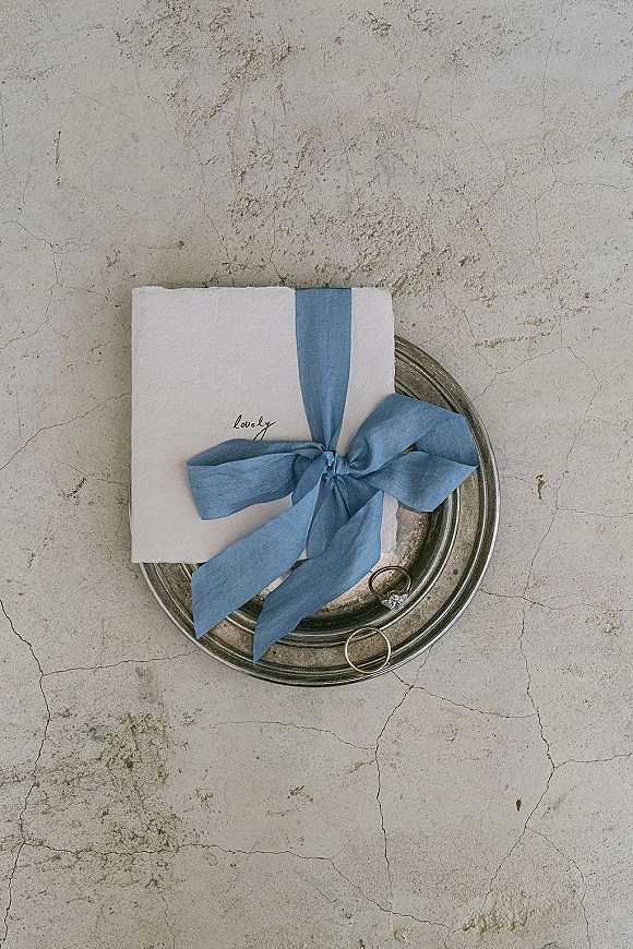 Invitation suite wedding invitation flat lay with handmade paper envelope, blue silk ribbon accent, and rings on a metal tray over stone surface