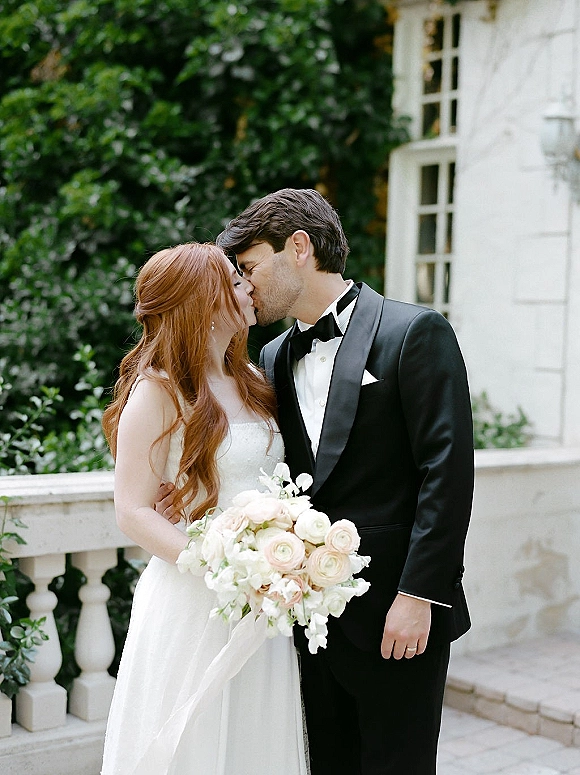 Wedding kiss portrait of bride and groom kissing, she holds a white and blush bouquet by a stone balustrade with garden greenery behind
