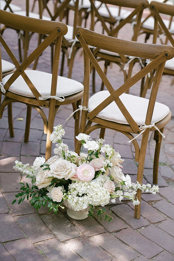 Ceremony seating with cross back chairs and white cushions, floral urn of roses and ranunculus lining an outdoor brick patio aisle