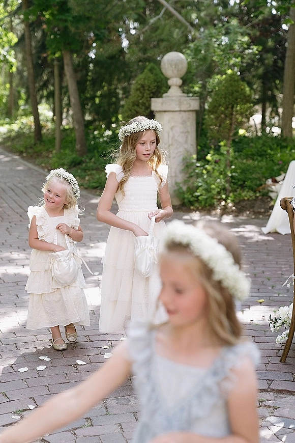 Flower girl walking down aisle in an ivory dress and baby’s breath flower crown, holding a petal pouch on a brick garden walkway