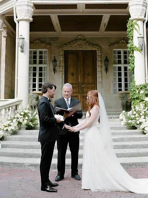Wedding ceremony with couple exchanging vows while holding hands, bride in lace dress and veil on manor house stone steps with florals