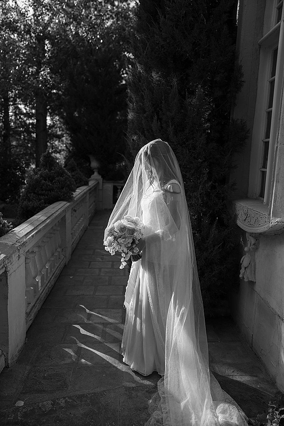 Bridal portrait in black and white of a bride in veil holding a bouquet on a stone walkway by a balustrade in garden light