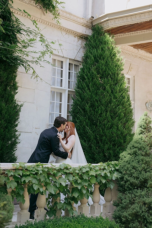 Wedding kiss portrait of bride and groom kissing on an ivy-covered stone balcony, her long veil and strapless dress beside his black tuxedo