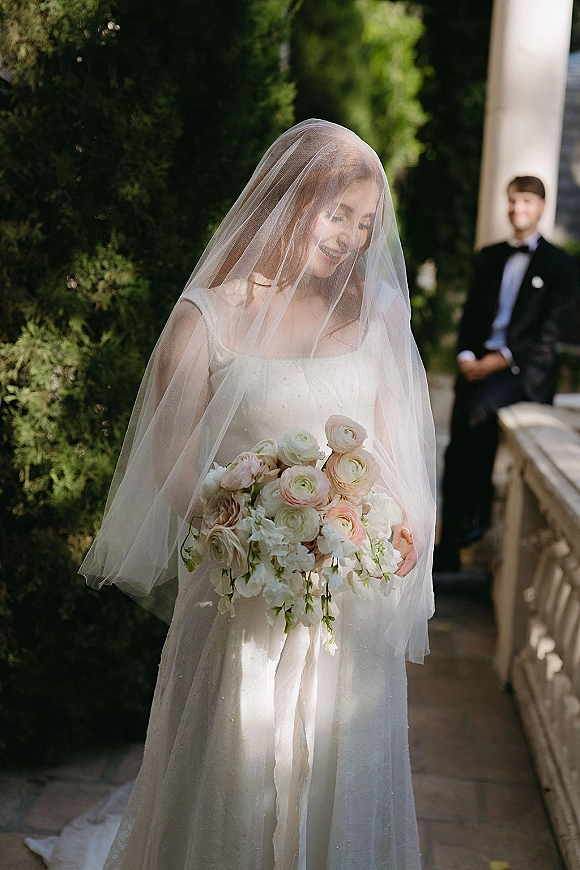 Bridal portrait of a bride holding bouquet with blush roses and greenery, veil over her face beside groom in tux by garden stone wall