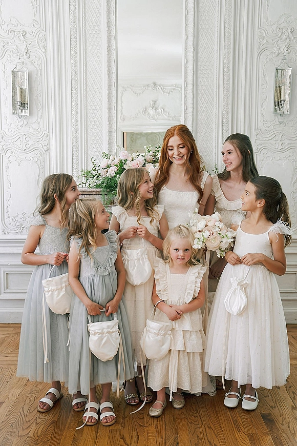 Bride with flower girls posing in flower girl dresses, holding blush rose bouquet and baskets before an ornate white wall and mirror