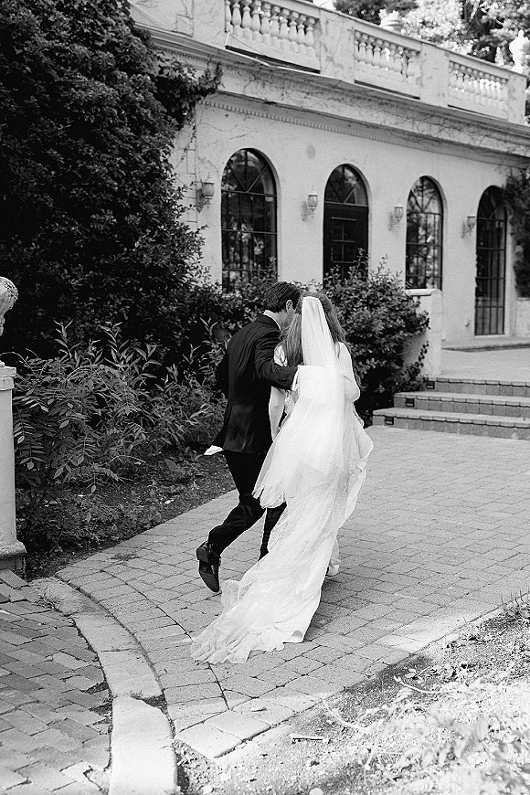 Wedding couple portrait in black and white, bride and groom walking away with cathedral veil flowing beside a stone building and garden path