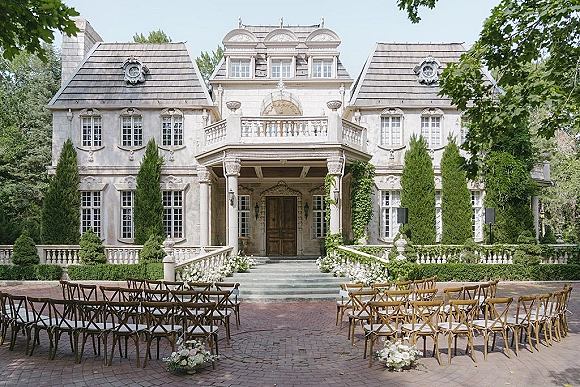Outdoor ceremony setup with estate wedding ceremony crossback chairs flanking a wide aisle of white flowers and greenery before a stone mansion facade
