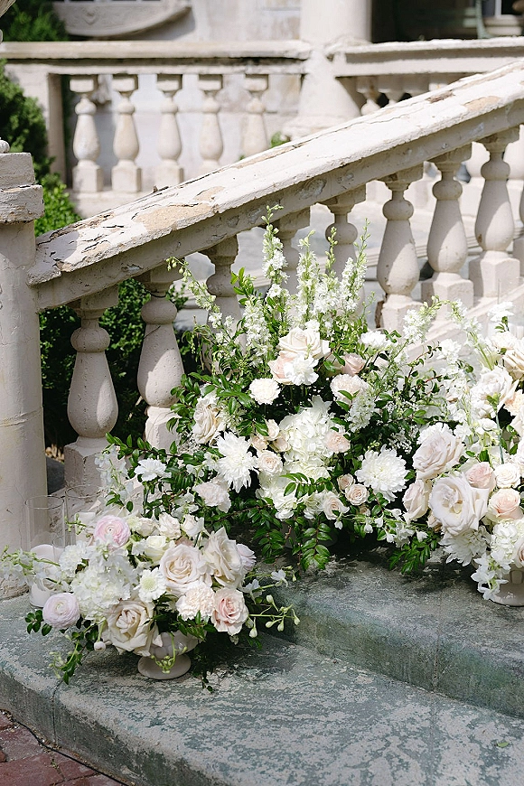 Wedding floral arrangement of white and blush roses with greenery and glass cylinder vases on a stone staircase by garden shrubs