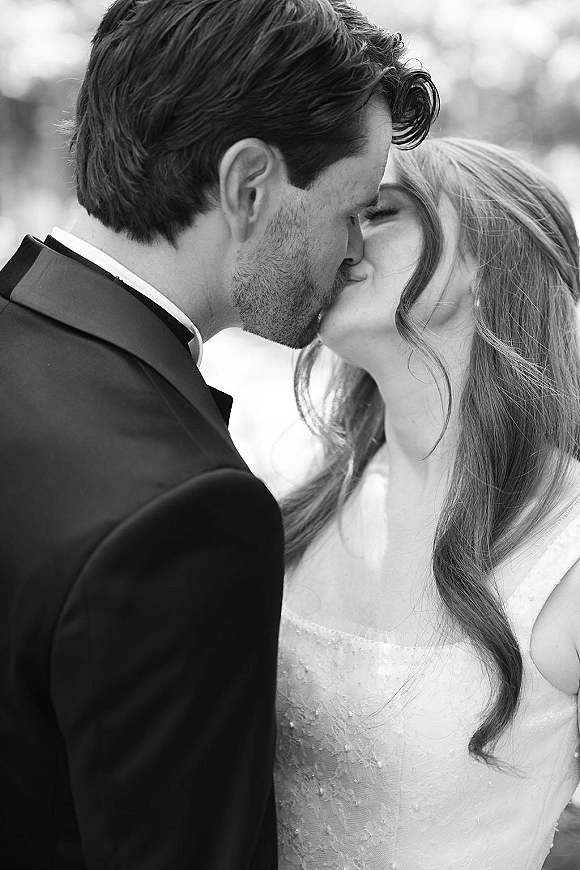 Wedding kiss portrait of the bride and groom kissing in a black tuxedo and lace bodice gown against softly blurred outdoor greenery